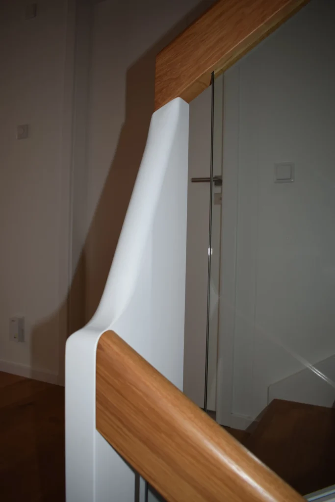 Nahaufnahme einer modernen Treppe mit einem weißen, glatten Geländer und einem hölzernen Handlauf vor einer schlichten Wand und einer Tür im Hintergrund.