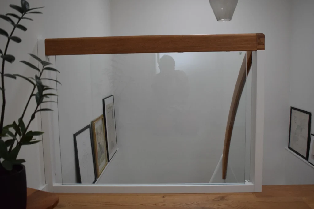 Ein gläsernes Treppengeländer mit einem hölzernen Handlauf, daneben eine Pflanze und mehrere gerahmte Bilder, die an der Treppenhauswand lehnen. Eine schwache Reflexion einer Person, die das Foto macht, ist im Glas sichtbar.