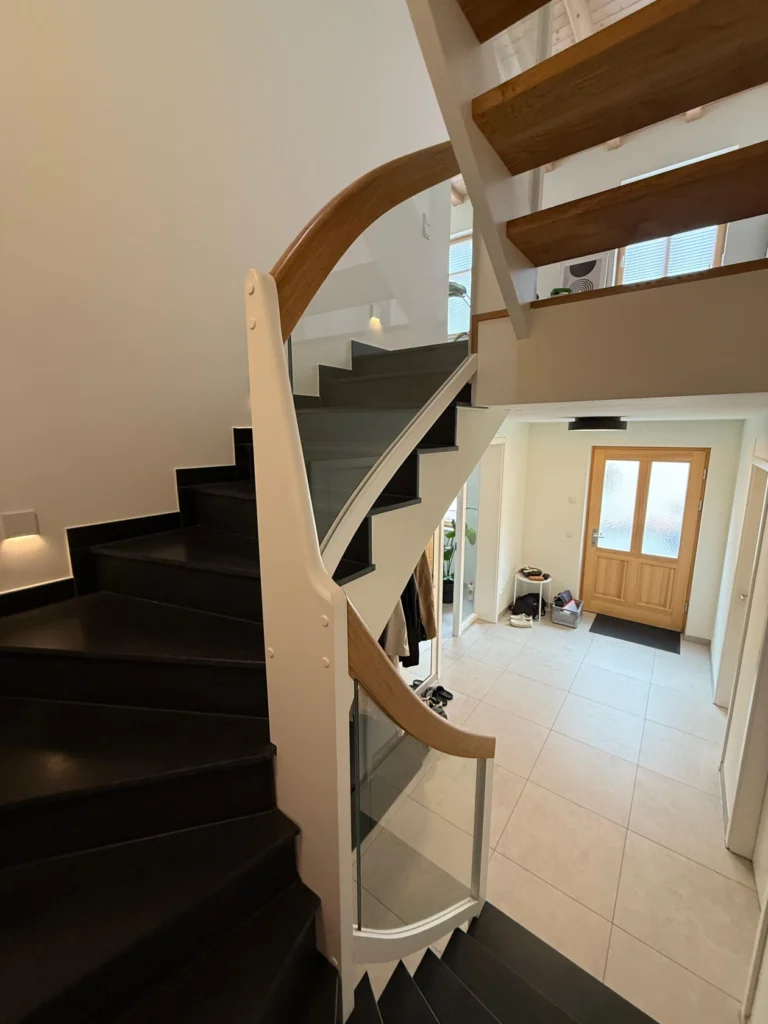 Ein modernes Wohnhaus mit einer Holztreppe, einem Glasgeländer und einem beigen Kachelboden. Die hölzerne Eingangstür lässt natürliches Licht herein. Schuhe und ein kleiner Tisch stehen neben dem Eingang.