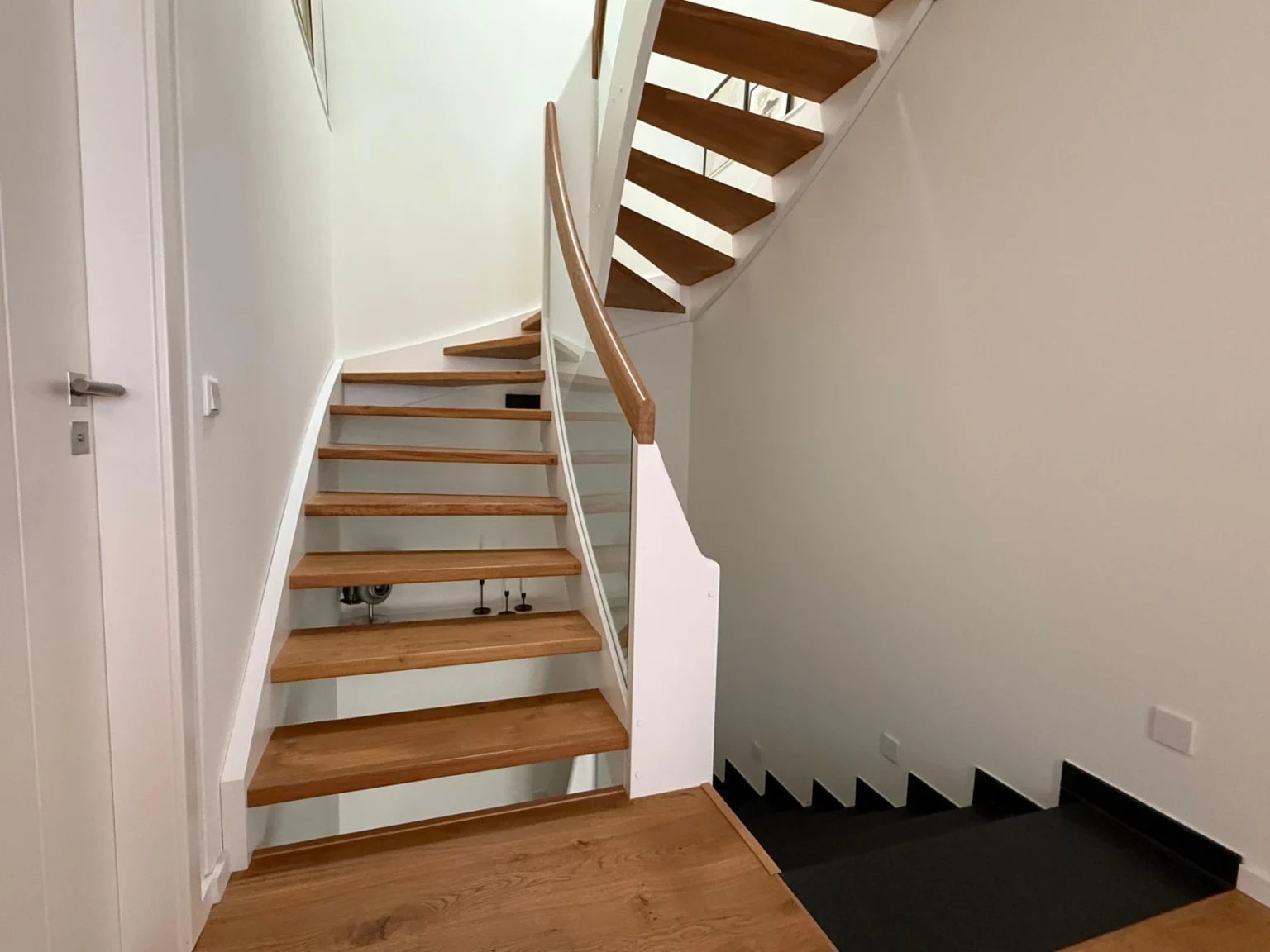 Holztreppe mit geschwungenem Handlauf und weißen Wänden, die zu einem Treppenabsatz und zu einer unteren Ebene mit dunklen Stufen führt. Der Raum ist modern und gut beleuchtet mit minimalem Dekor.