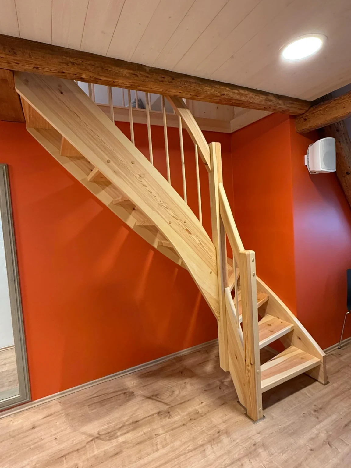 Eine Holztreppe mit Handlauf und vertikalen Geländerstäben steht vor einer orangefarbenen Wand in einem Raum mit Holzboden und freiliegenden Holzbalken an der Decke.