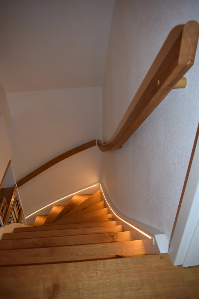 Gebogene Holztreppe mit Handlauf an einer weißen Wand, beleuchtet durch LED-Leuchtbänder entlang der Kante jeder Stufe, Blick von oben nach unten.