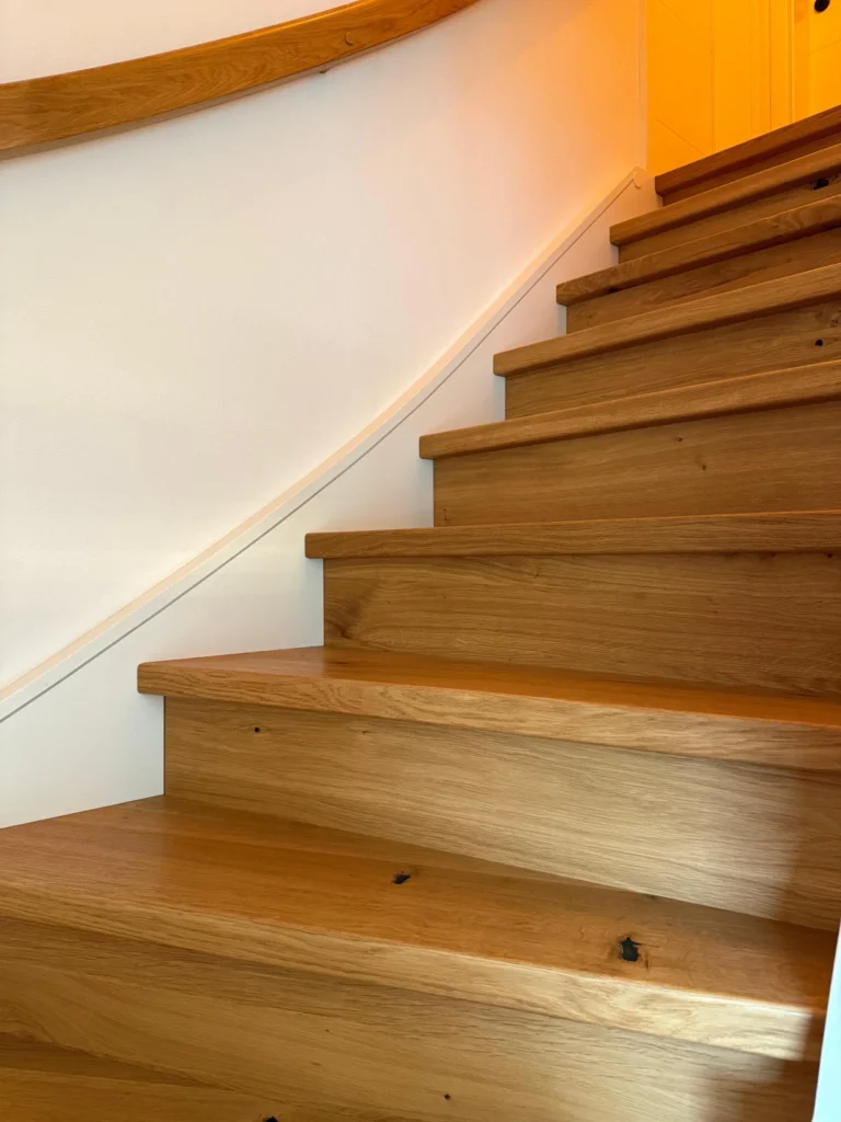 Nahaufnahme einer modernen Holztreppe mit einer weißen Wand und einem hölzernen Handlauf auf der linken Seite, die zu einer warm beleuchteten Tür hinaufführt.