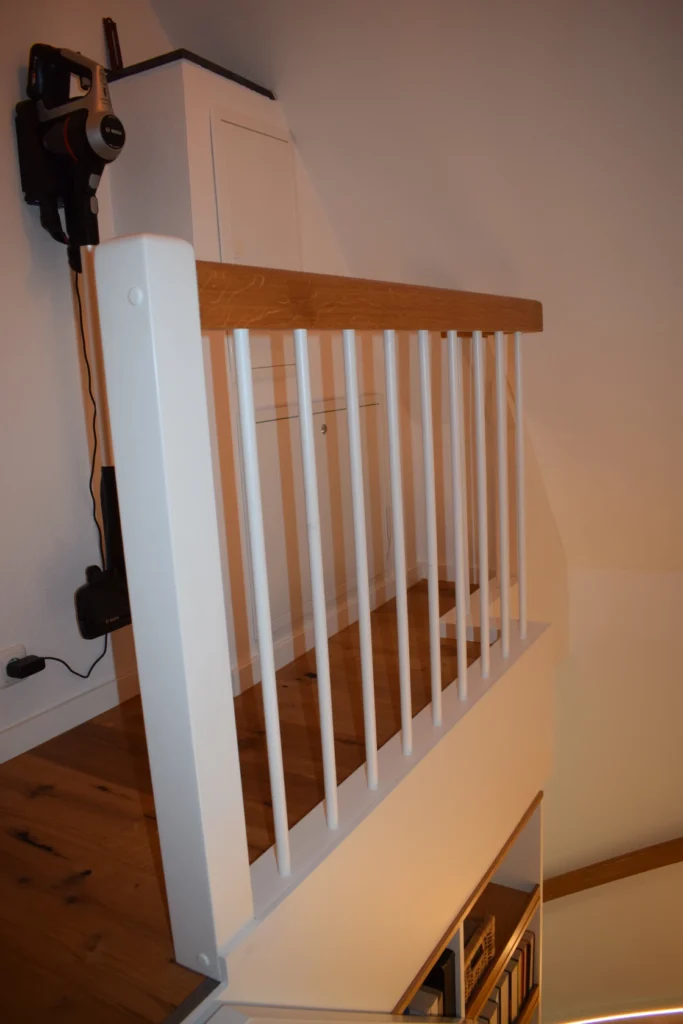 Ein hölzernes Treppenpodest mit weißem vertikalem Geländer und hellbraunem Handlauf, angrenzend an eine weiße Wand mit einer montierten Ladestation und einem daran hängenden Staubsauger.