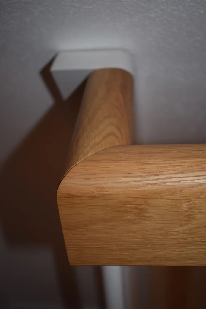 Nahaufnahme eines hölzernen Handlaufs mit glatten, abgerundeten Kanten, der an einer weißen Wandhalterung an einer hellen Wand befestigt ist und einen deutlichen Schatten auf die Oberfläche wirft.