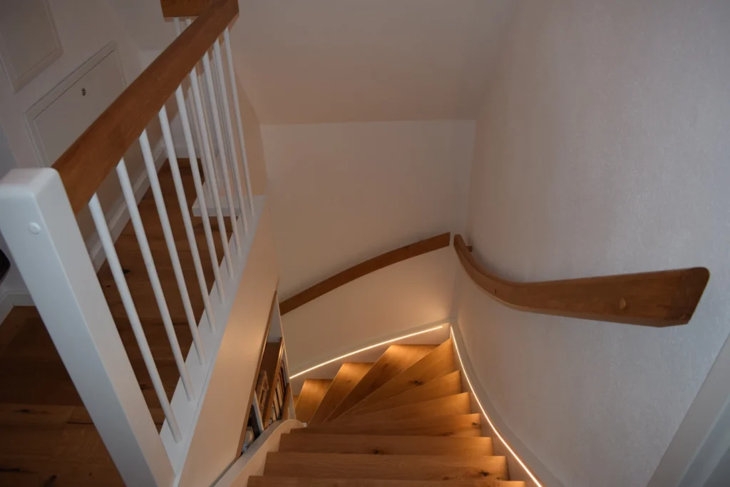 Eine Holztreppe mit weißem Geländer schlängelt sich nach unten, mit eingebauter LED-Beleuchtung entlang der Stufen und einem hölzernen Handlauf, der an einer weißen Wand befestigt ist.
