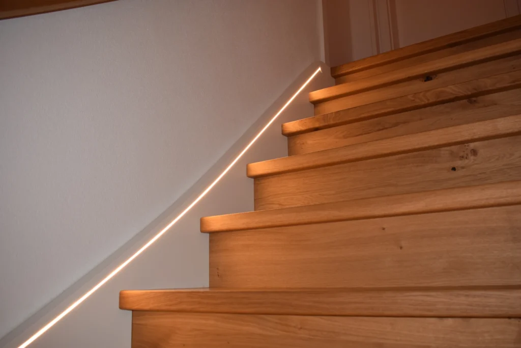 Holztreppe mit eingebauter LED-Beleuchtung entlang der Wand, die die Stufen in einem weichen, warmen Licht erstrahlen lässt. Die Treppe hat glatte Holzstufen und Setzstufen, und die Beleuchtung sorgt für einen modernen und eleganten Look.