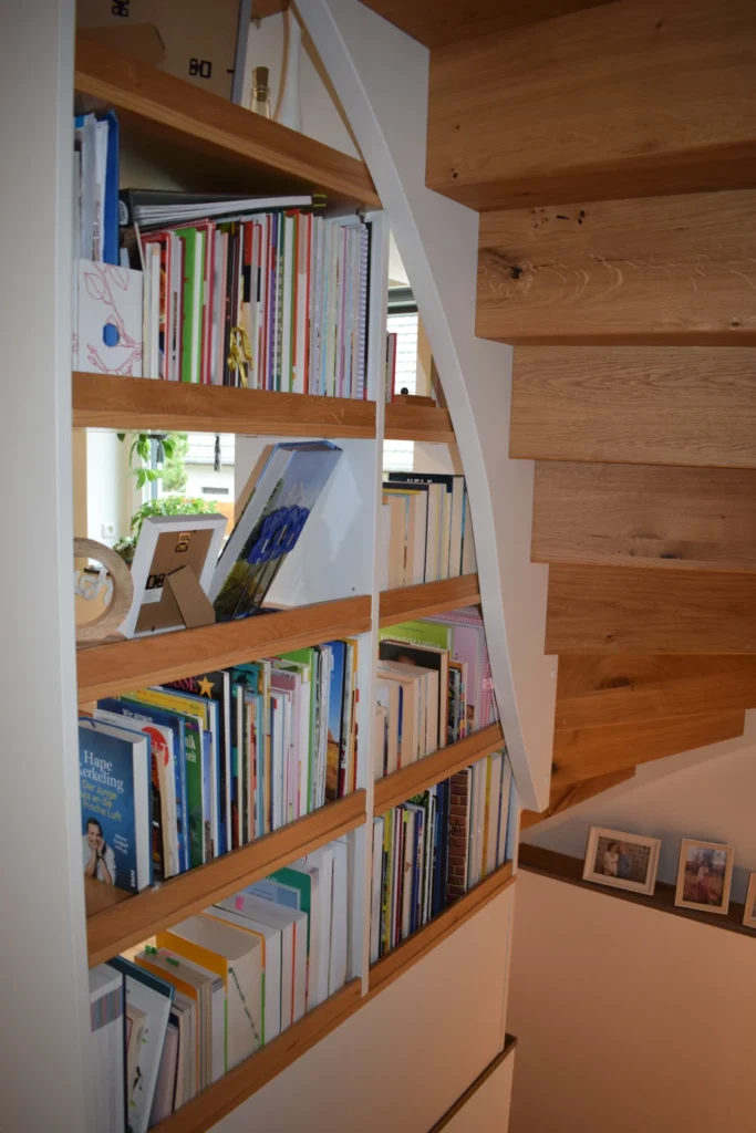 Ein mit verschiedenen Büchern gefülltes Bücherregal ist in die Wand unter einer Holztreppe eingebaut. Einige gerahmte Fotos sind auf dem benachbarten Sims ausgestellt, und ein Fenster lässt natürliches Licht herein.