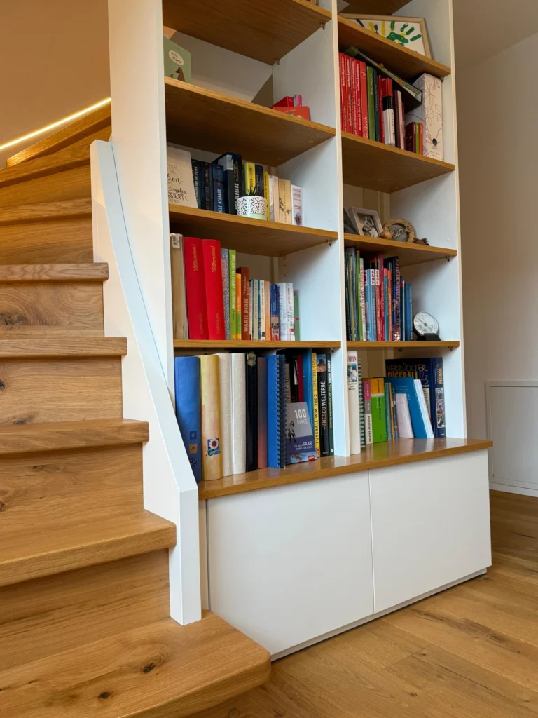 Eine Holztreppe neben einem hohen weißen Bücherregal, gefüllt mit bunten Büchern und einigen dekorativen Gegenständen, alles auf einem hellen Holzboden in einem hellen, modernen Interieur.