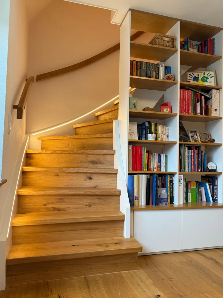 Eine Holztreppe mit eingebauter LED-Beleuchtung schlängelt sich neben einem hohen, modernen Bücherregal, das mit Büchern und Dekorationsartikeln gefüllt ist, nach oben. Der Boden und die Treppe sind aus passendem hellem Holz gefertigt.