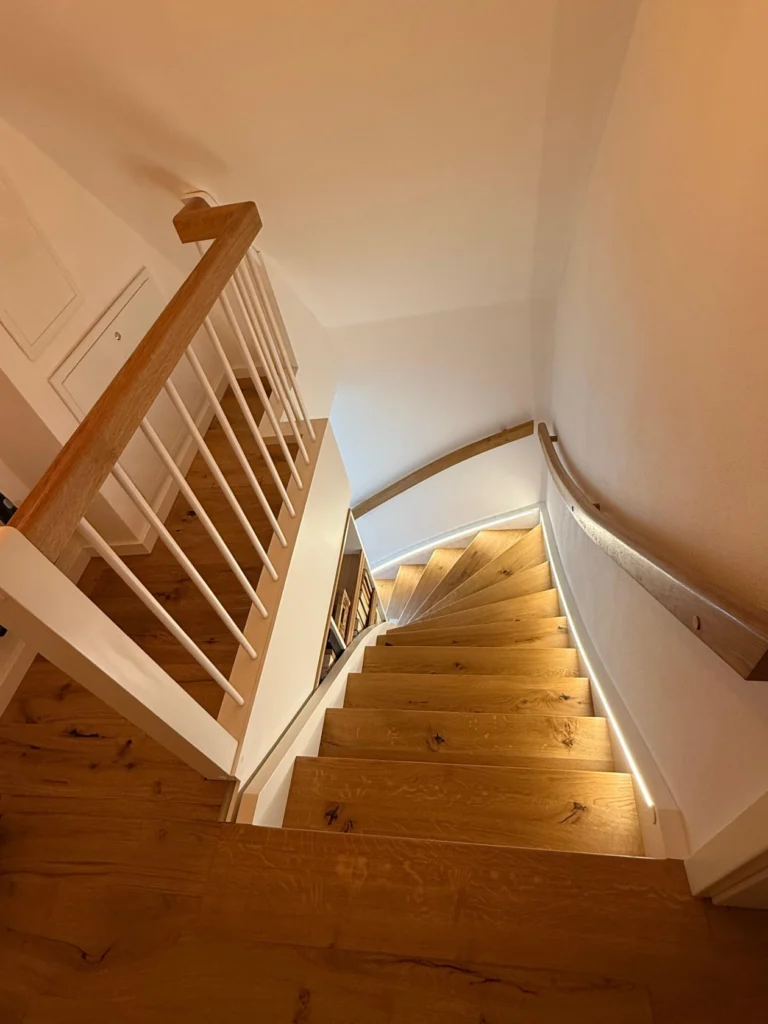 Blick von unten auf eine Holztreppe mit weißem Geländer und geschwungenem Handlauf, die von Leuchten entlang der Wand warm beleuchtet wird und zu einem unteren Stockwerk in einem modernen Interieur führt.