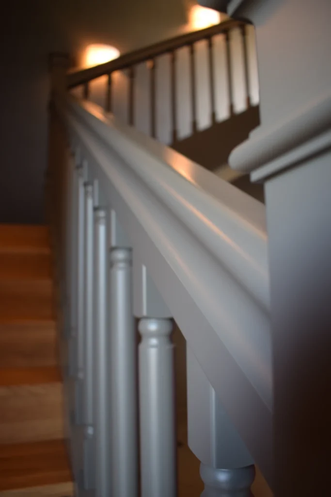 Nahaufnahme eines grauen Holztreppengeländers mit vertikalen Balustern, das in ein schwach beleuchtetes Obergeschoss führt. Warmes Licht beleuchtet sanft Teile der Stufen und des Geländers.