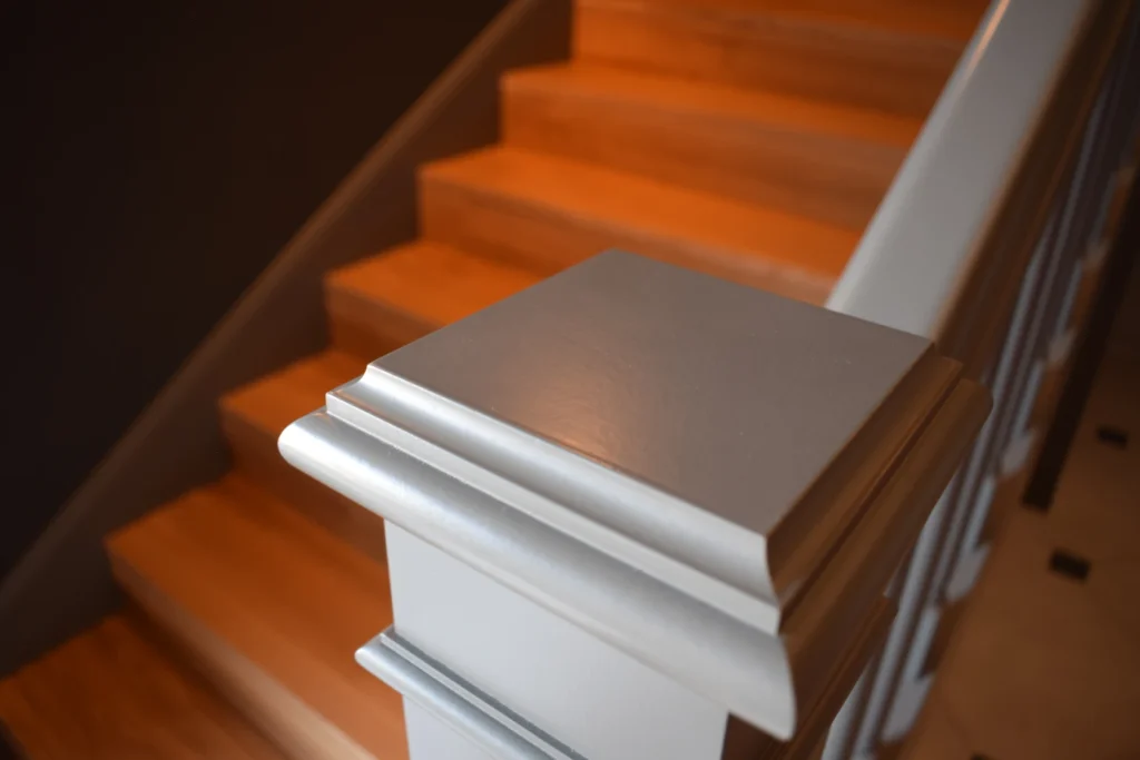 Nahaufnahme eines weißen hölzernen Treppengeländerpfostens mit nach oben führenden Holzstufen im Hintergrund, sanft beleuchtet, um die glatte Oberfläche und die warmen Holztöne hervorzuheben.