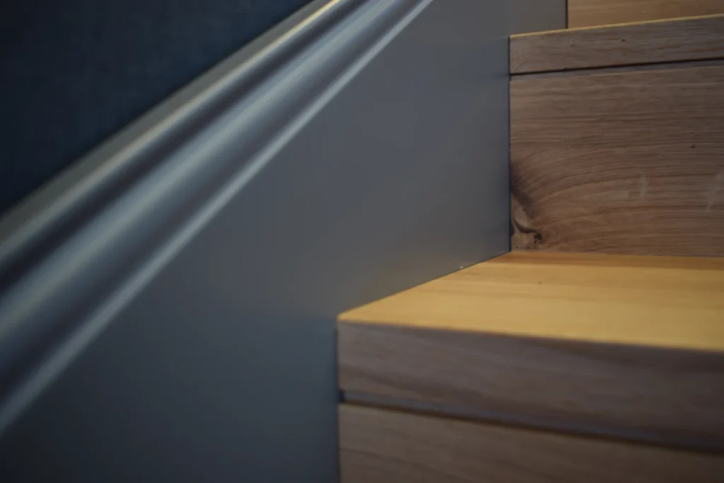Nahaufnahme einer Holztreppe mit einer glatten, grau gestrichenen Wand und einem Handlauf daneben, der die Ecke zeigt, in der die Stufen auf die Wand treffen.