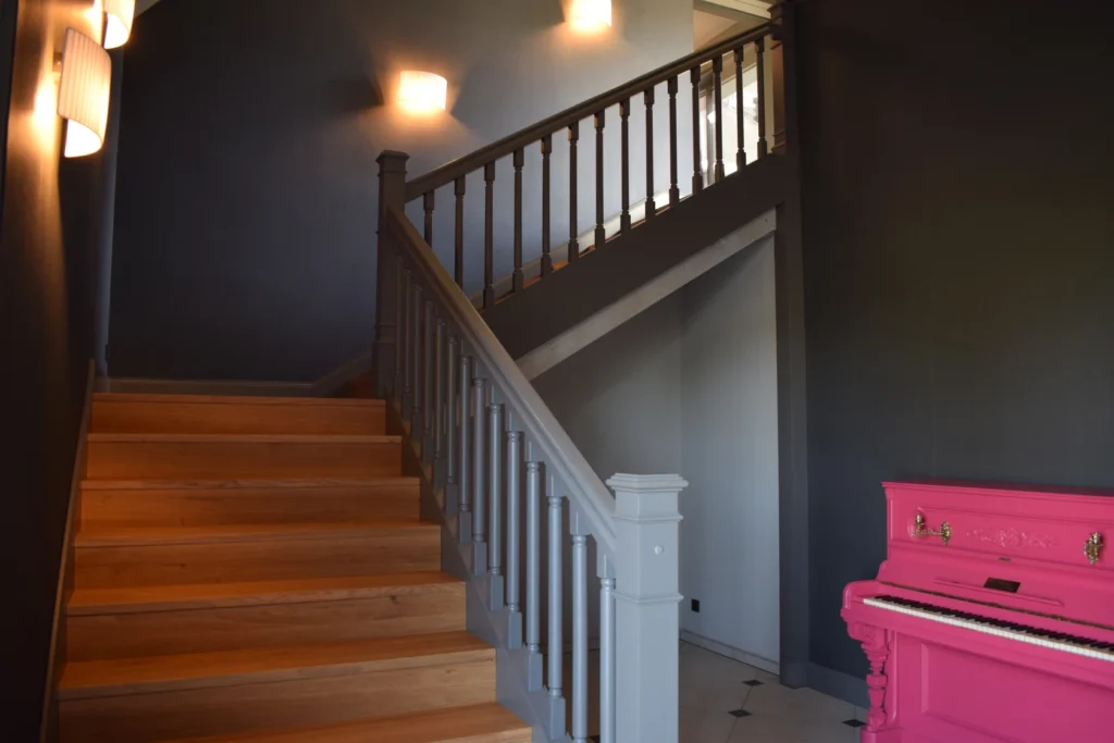 Eine Treppe mit hölzernen Stufen und grauem Geländer führt zu einem Treppenabsatz hinauf; sanfte Wandleuchten erhellen den Raum, und ein hellrosa Klavier steht auf der rechten Seite.