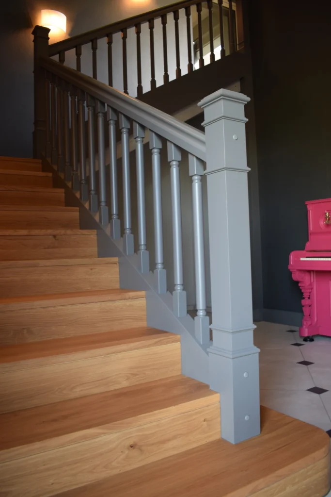 Eine Holztreppe mit grauem Geländer führt neben einem gefliesten Boden nach oben; an der dunklen Wand im Hintergrund steht ein hellrosa Klavier.