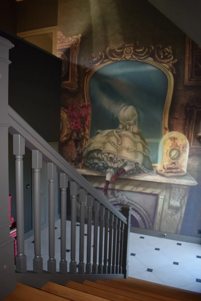 Eine Wandmalerei über einer Treppe zeigt ein Mädchen in einem Rüschenkleid, das an einem Frisiertisch sitzt, von hinten gesehen, mit einem verzierten Rahmen und Gegenständen auf dem Tisch; im Vordergrund ein graues Treppengeländer.