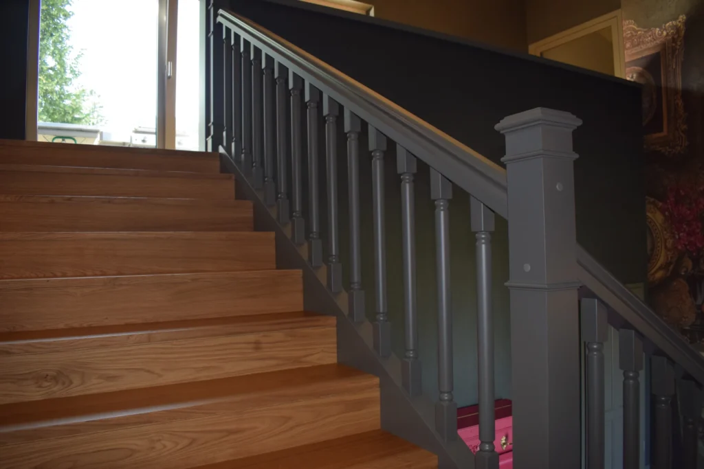 Eine Holztreppe mit breiten Stufen und einem dunkelgrauen Geländer, die zu einem hellen Fenster führt. Unter der Treppe befinden sich zwei rosa Aufbewahrungsboxen. Die Umgebung scheint ein Innenraum zu sein, in den natürliches Licht fällt.