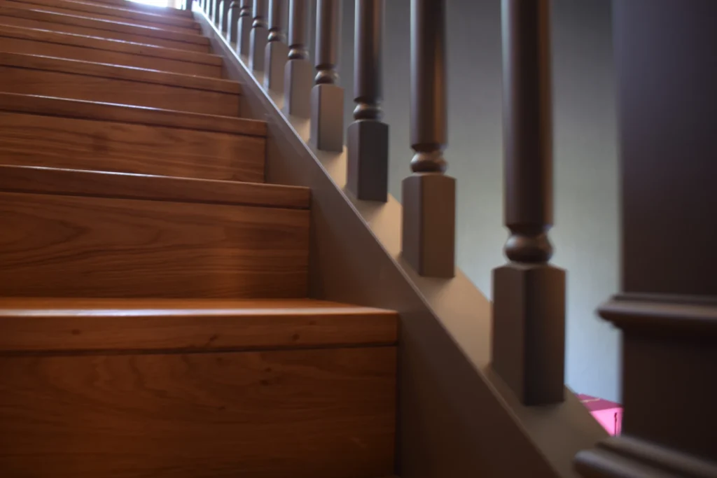 Nahaufnahme einer Holztreppe mit braun polierten Stufen und dunklen Geländerstäben, die nach oben führt. Sanftes Licht erhellt die Treppe und schafft eine warme und einladende Atmosphäre.