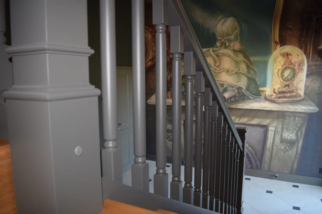 Eine Treppe mit dunkelgrauen Geländerstäben und einem Wandgemälde im Hintergrund, das eine Person zeigt, die mit dem Rücken zugewandt an einem Tisch mit einer verschnörkelten Uhr sitzt.