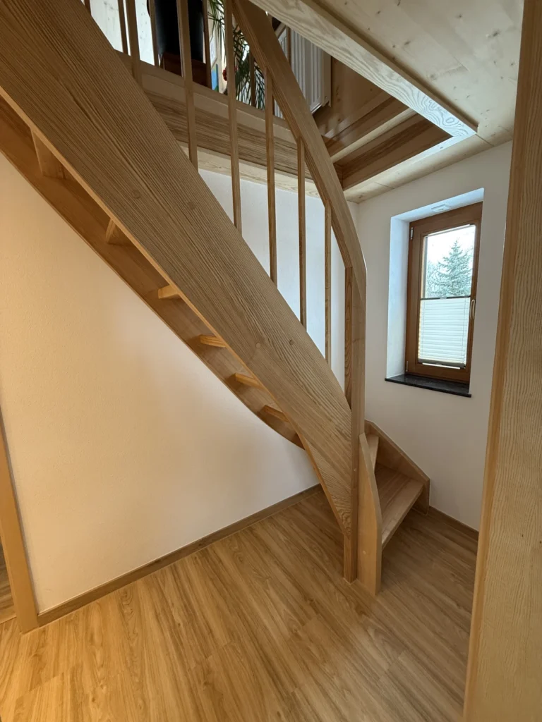 Holztreppe mit vertikalem Geländer in einem hellen Raum mit hellen Holzböden und Wänden. Ein kleines Fenster mit einem Holzrahmen lässt natürliches Licht herein. Das Design zeichnet sich durch klare, moderne Linien aus.