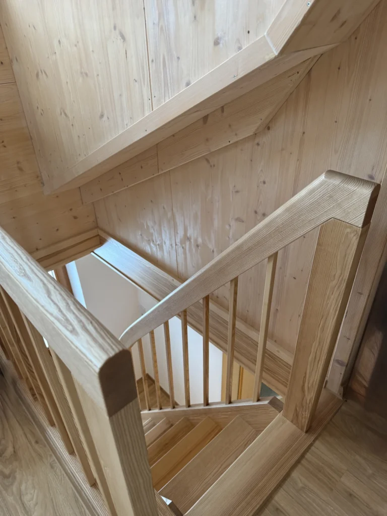 Eine Holztreppe mit vertikalen Geländern und glatten Handläufen, von oben gesehen. Die Treppe und die sie umgebenden Wände sind aus hellem Holz gefertigt, was dem Raum ein warmes, natürliches Aussehen verleiht.