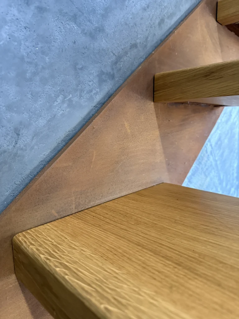 Nahaufnahme einer modernen Holztreppe mit einer Betonwand und einem Metallakzent unter den Stufen, der die bei der Gestaltung der Treppe verwendeten Texturen und Materialien hervorhebt.