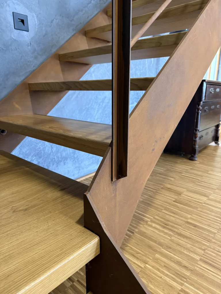 Nahaufnahme einer modernen Treppe mit Holzstufen und einer seitlichen Metallstütze vor einer strukturierten grauen Wand. Im Hintergrund ist ein Teil einer dunklen Holzkommode zu sehen, zusammen mit einem holzgemusterten Bodenbelag.