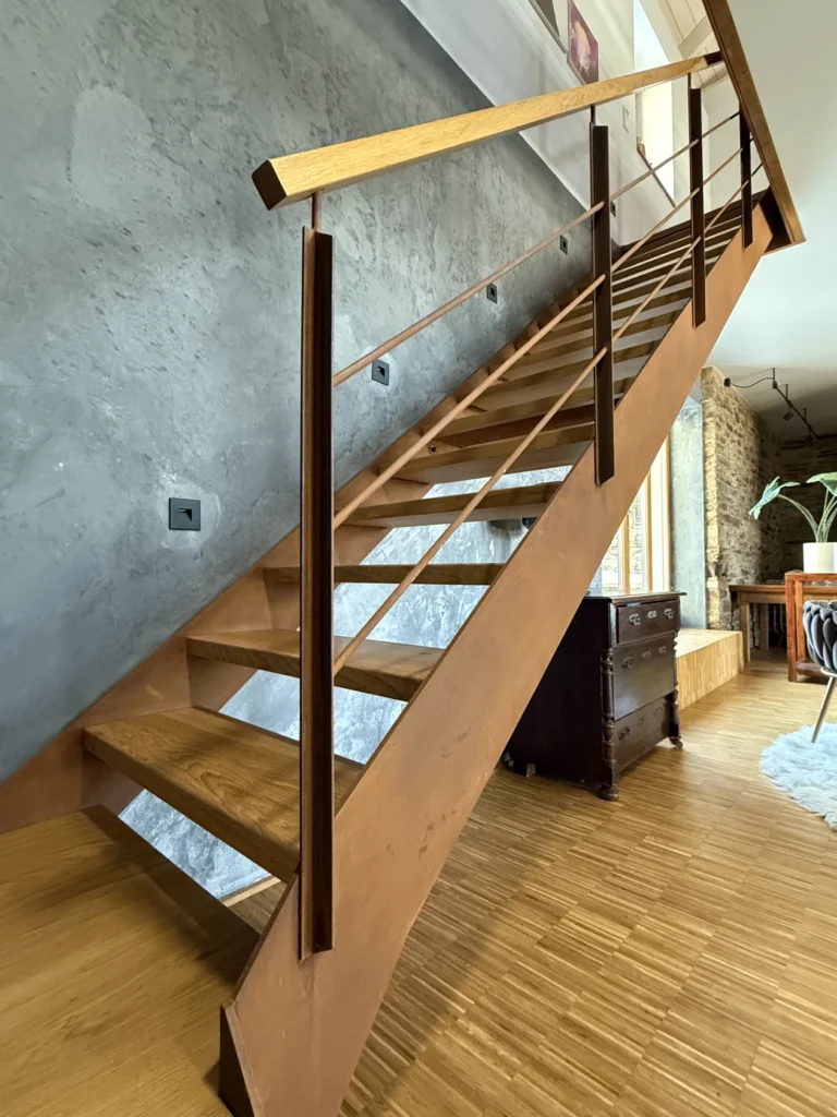 Eine moderne Holztreppe mit Metallgeländer an einer strukturierten grauen Wand; die Stufen führen in ein Obergeschoss in einem stilvollen, gut beleuchteten Raum mit Holzböden und zeitgenössischem Dekor.