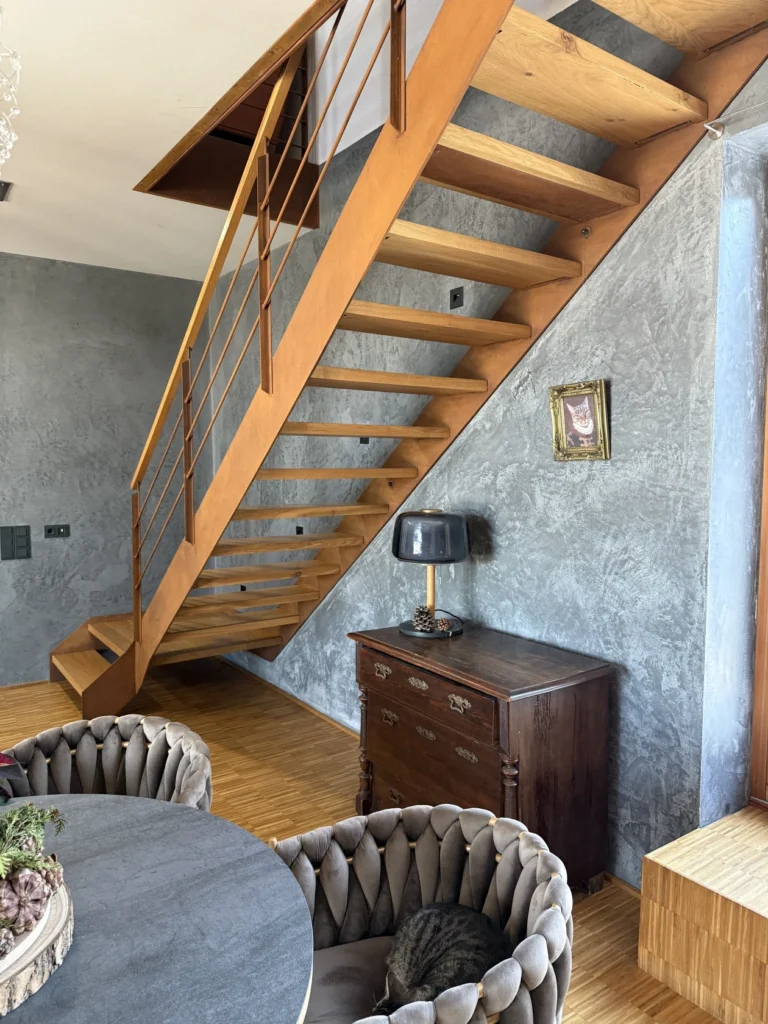 Eine moderne Holztreppe mit offenen Setzstufen steht in einem sonnendurchfluteten Raum mit grau strukturierten Wänden. Unter der Treppe stehen eine alte Holztruhe, eine Lampe und ein gerahmtes Bild. Eine Katze sitzt zusammengerollt auf einem Plüschsessel neben einem runden Tisch.