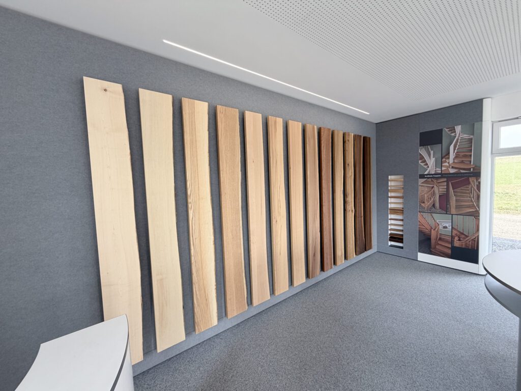 In einem modernen Ausstellungsraum sind an einer grauen Wand mehrere vertikale Holzbretter in verschiedenen Farbtönen angebracht, während im Hintergrund Wandtafeln mit Fotos von Holzarbeiten zu sehen sind. Der Raum ist mit Teppichboden ausgelegt und hell und gleichmäßig beleuchtet.