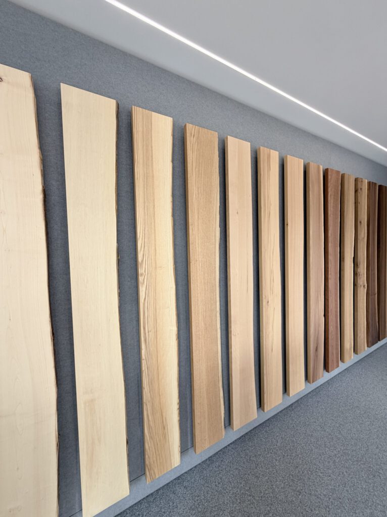 Mehrere vertikale Holzbretter in verschiedenen Farben und Maserungen sind nebeneinander an einer grauen Wand angebracht, mit einer weißen Decke und Lichtbändern darüber. Die Dielen reichen von hellen bis zu dunklen Tönen.