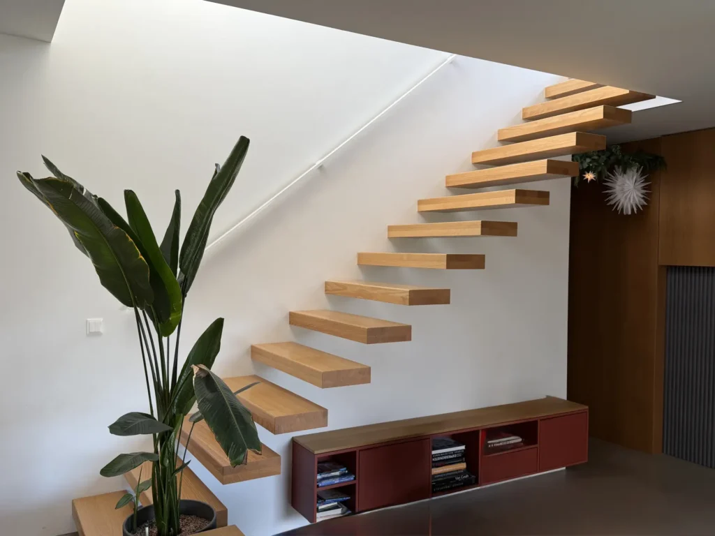 Schwebende Holztreppe mit dicken, rechteckigen Stufen an einer weißen Wand, über einem modernen roten Schrank mit Büchern und einer Topfpflanze in einem hellen, minimalistischen Interieur.