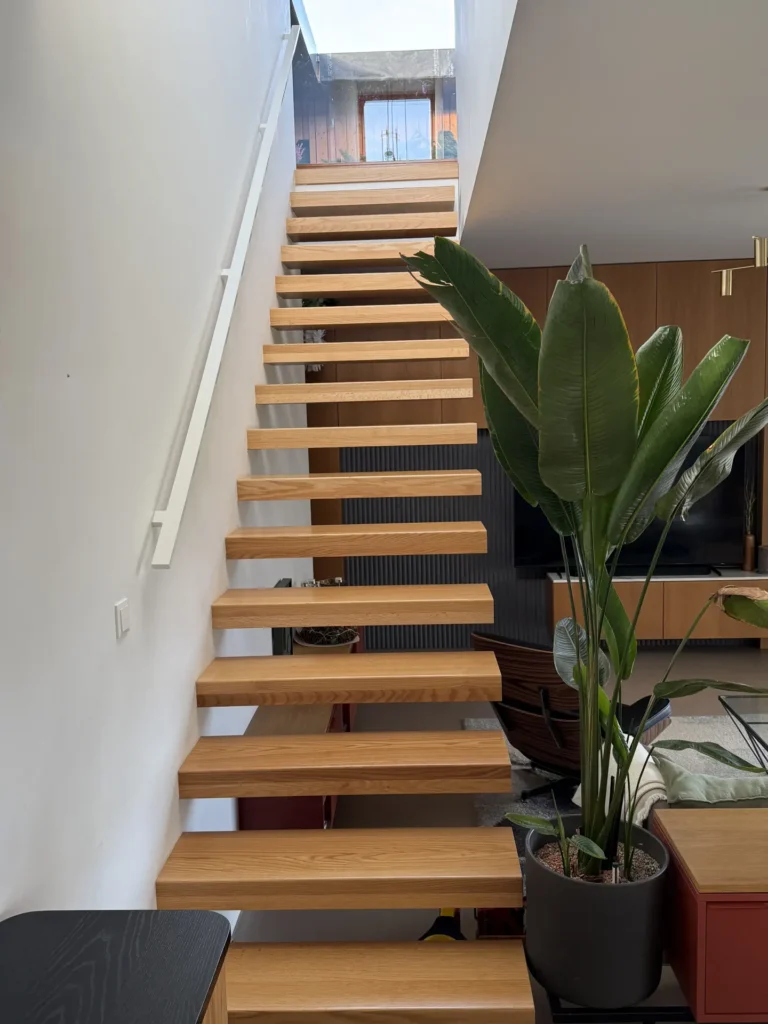 Eine moderne schwebende Holztreppe mit offenen Setzstufen führt entlang einer weißen Wand nach oben. Am Fußende steht eine große grüne Zimmerpflanze neben der Treppe in einem modernen Wohnraum mit natürlichem Licht.