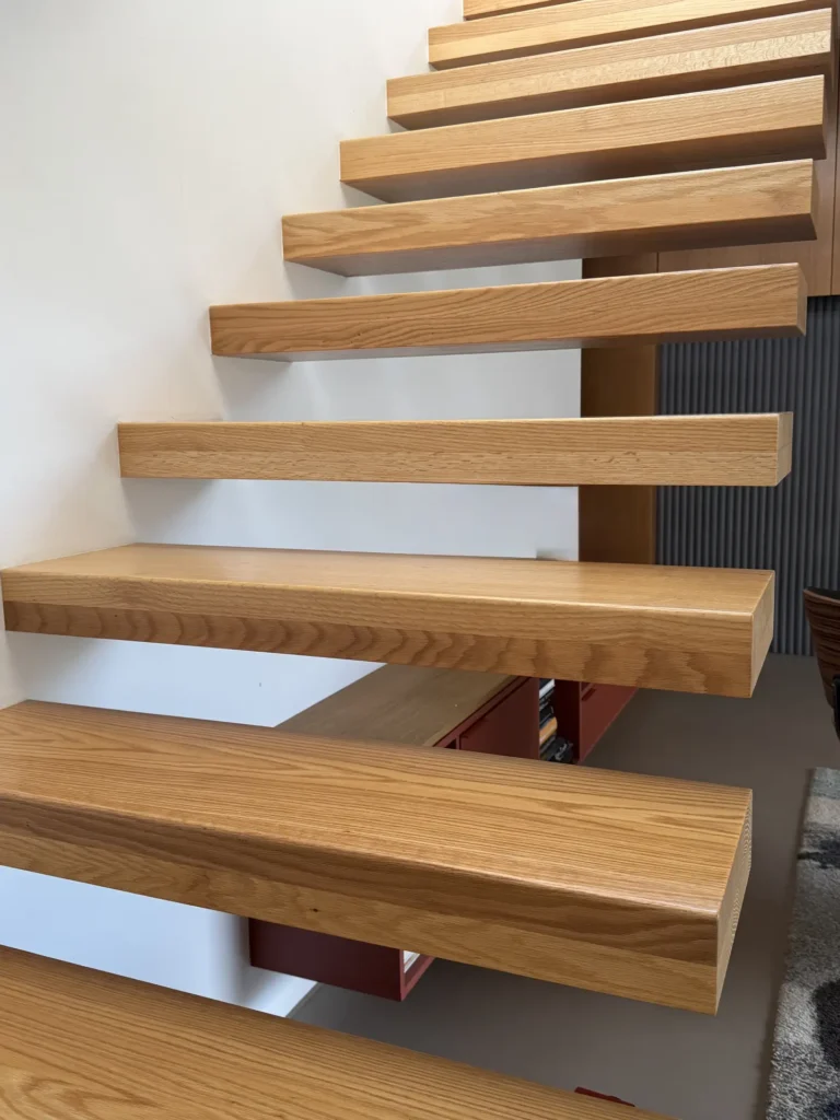 Schwebende Holztreppe mit dicken rechteckigen Stufen, die an einer weißen Wand befestigt sind. Es gibt keine sichtbare Stütze darunter, wodurch ein modernes, minimalistisches Aussehen entsteht. Darunter sind ein Teil eines Teppichs und ein Regal zu sehen.