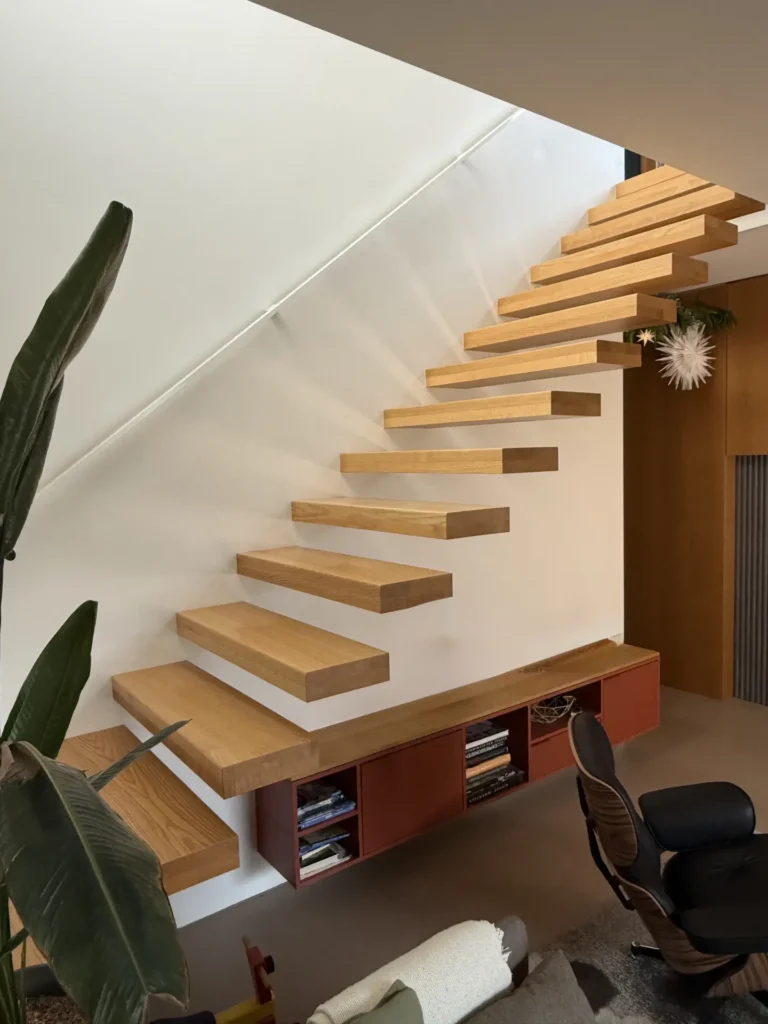 Schwebende Holztreppe mit dicken, rechteckigen Stufen, die an einer weißen Wand befestigt ist, über einem eingebauten Regal mit Büchern. Ein moderner Stuhl, eine große Pflanze und ein minimalistisches Dekor sind in diesem modernen Raum zu sehen.