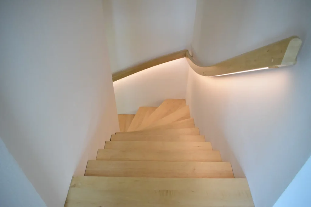 Blick von unten auf eine hölzerne Wendeltreppe mit hellen Stufen, weißen Wänden und einem geschwungenen hölzernen Handlauf mit eingebauter Beleuchtung auf der rechten Seite.