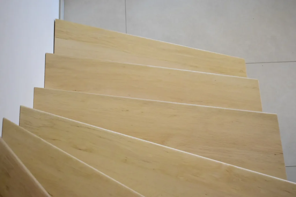 Blick von oben auf eine helle Holztreppe mit modernem, minimalem Design, die sich von einem hellen Fliesenboden abhebt.