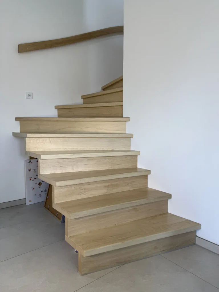 Eine moderne Holztreppe ohne Geländer schlängelt sich vor einer weißen Wand in einem minimalistischen Interieur nach oben. Die Stufen sind breit und aus hellem Holz, und ein Holzbalken ist an der Wand über der Treppe angebracht.