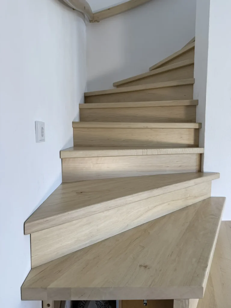 Eine helle Holztreppe mit schrägen, unregelmäßig geformten Stufen schlängelt sich entlang einer weißen Wand mit einer sichtbaren Steckdose nach oben. Die Stufen erscheinen schmal und ungleichmäßig breit.