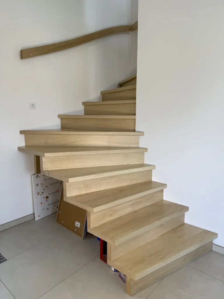 Eine moderne Holztreppe mit offenen Setzstufen und minimalistischem Design schlängelt sich entlang einer weißen Wand nach oben, wobei unter den ersten Stufen einige Bücher und Gegenstände aufbewahrt werden.