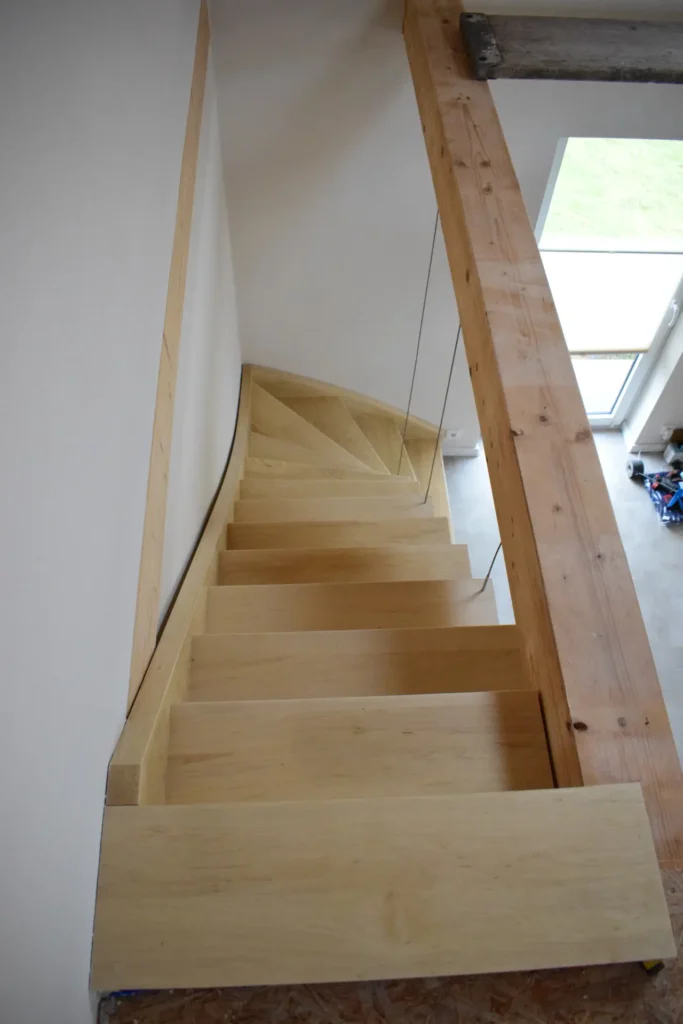 Blick von oben auf eine moderne, helle Holztreppe mit einer Drehung am unteren Ende, die entlang einer weißen Wand und eines Holzbalkens in einen hellen Raum mit großen Fenstern hinabführt.