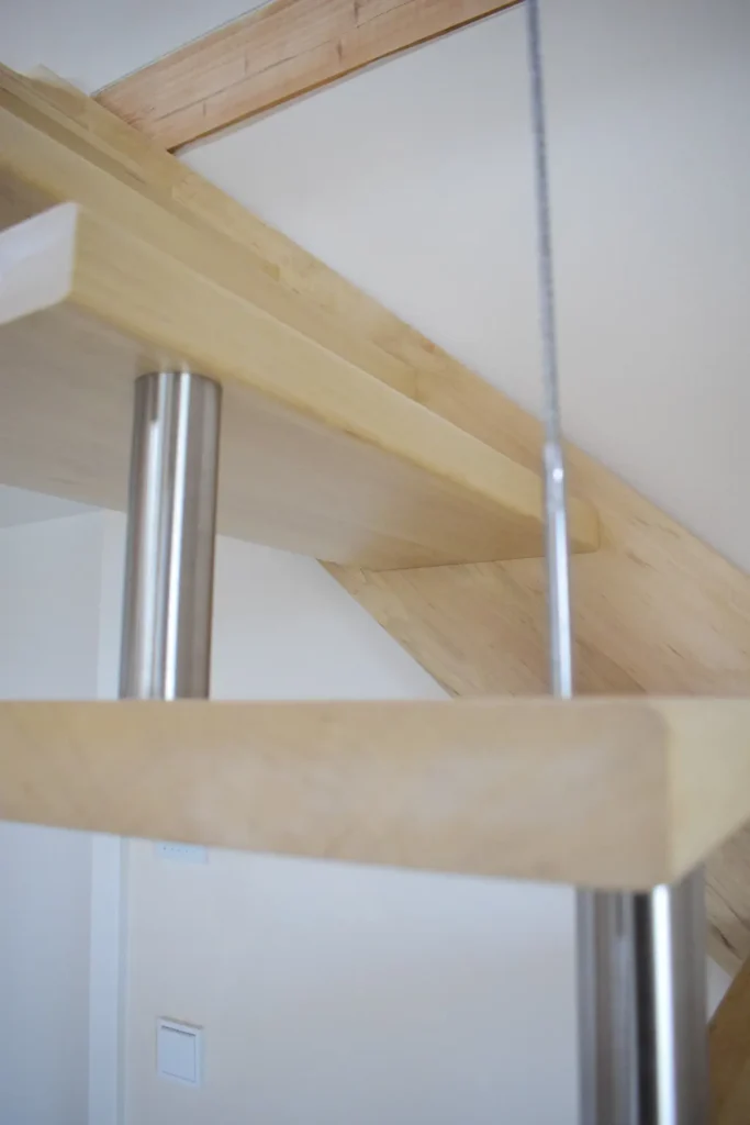 Nahaufnahme einer modernen Treppe mit hellen Holzstufen, die von zylindrischen Metallstäben und einem Kabel getragen werden, vor einem weißen Wandhintergrund.