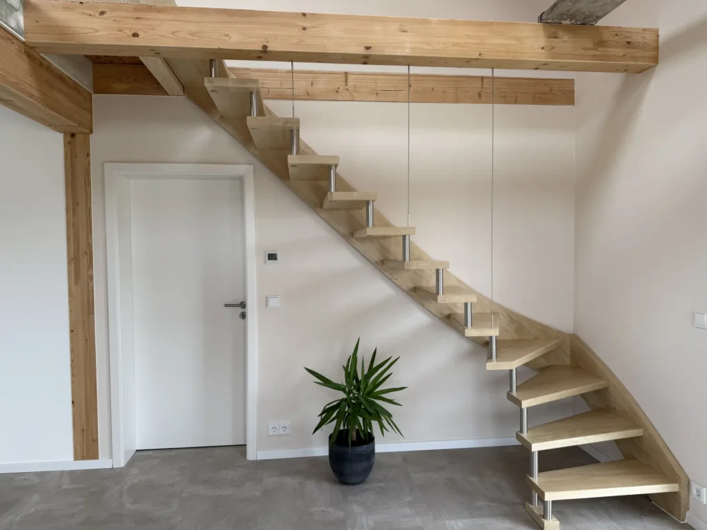 Eine moderne, schwebende Holztreppe mit offenen Setzstufen schlängelt sich an einer weißen Wand nach oben. Am Fuß der Treppe steht eine grüne Topfpflanze, links befindet sich eine weiße Tür. Der Raum hat freiliegende Holzbalken und ein minimalistisches Design.