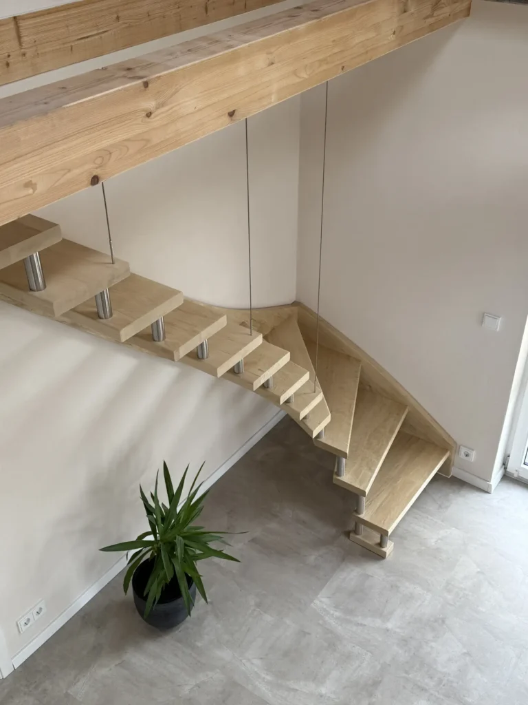 Eine moderne schwebende Holztreppe mit Metallstützen, die an einer weißen Wand befestigt ist, führt von einem Loftbereich zu einem hellgrauen Boden. Am Fuß der Treppe steht eine grüne Topfpflanze in einem schwarzen Übertopf. Der Raum ist minimalistisch und hell.