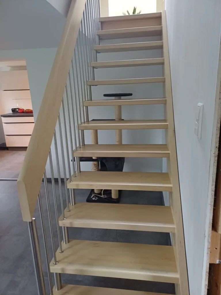 Eine helle Holztreppe mit offenen Setzstufen und Metallgeländer. Unter der Treppe steht ein Kratzbaum mit einer schwarzen Katze auf der unteren Ebene, deren Augen unter den Stufen sichtbar sind. Im Hintergrund ist eine Küche zu sehen.