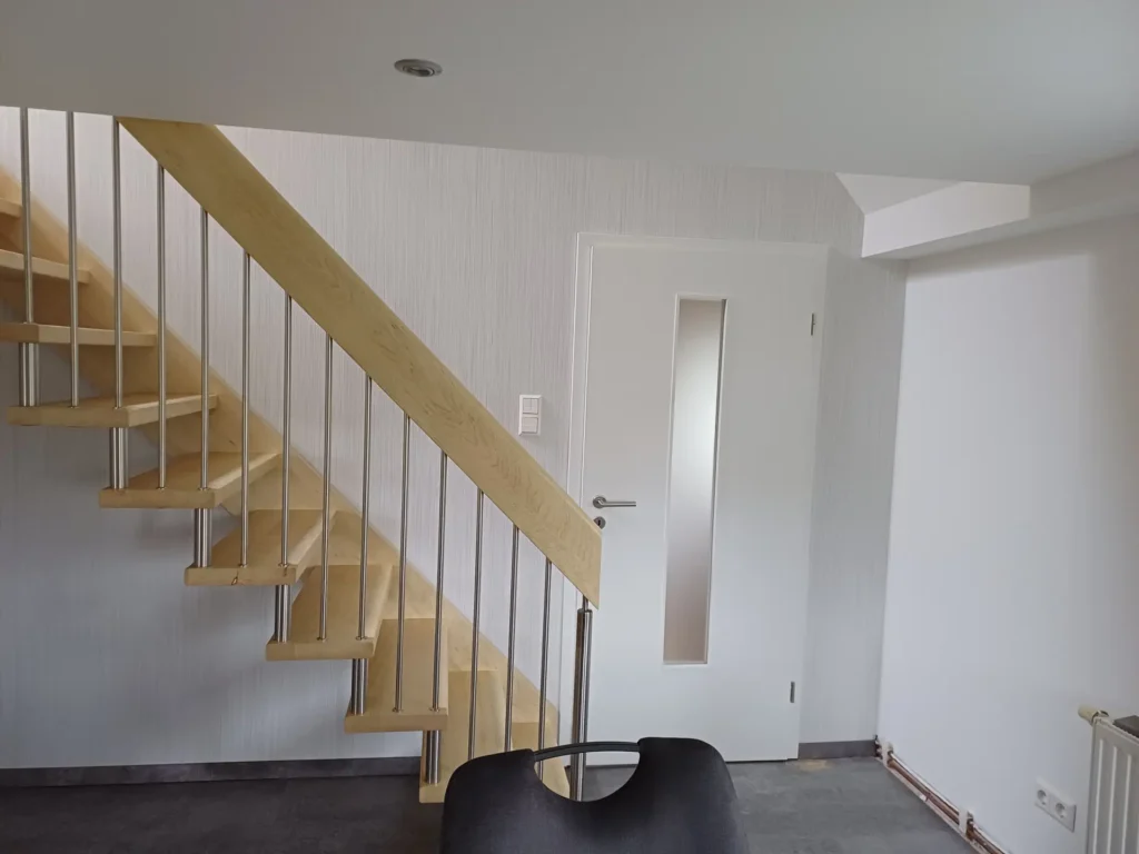 Ein minimalistisches Interieur mit einer hellen Holztreppe mit Metallgeländer, einer weißen Tür mit einer vertikalen Glasscheibe, weißen Wänden und einem schwarzen Stuhl im Vordergrund.