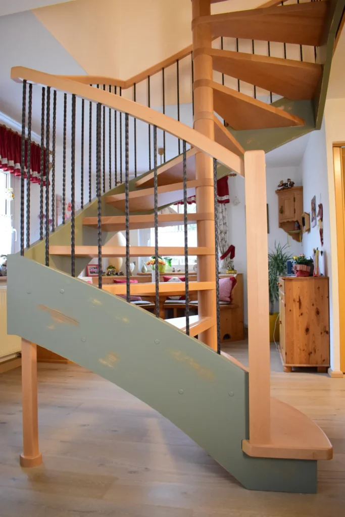 Eine hölzerne Wendeltreppe mit hellen und natürlichen Holztönen, schwarzen Metallgeländerstäben und einem grün-blau gestrichenen Sockel in einem gemütlichen, gut beleuchteten Wohnraum mit Holzmöbeln und Dekorationsartikeln.