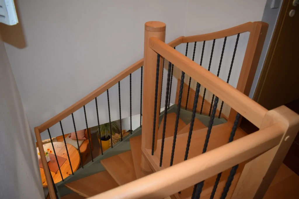 Eine hölzerne Wendeltreppe mit schwarzen Metallgeländerstäben, von oben gesehen. Unten sind ein kleiner Tisch mit einer orangefarbenen Tischdecke, eine Topfpflanze und ein Stuhl zu sehen.