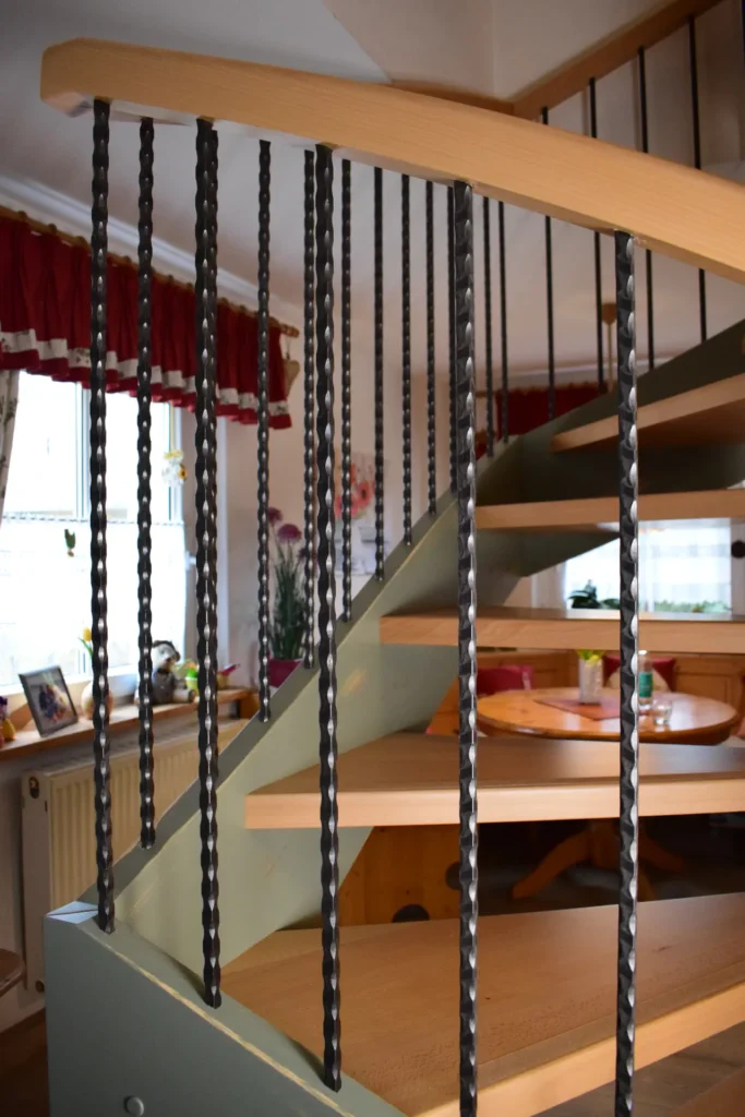 Holztreppe mit schwarzen, gedrehten Metallgeländern in einem gemütlichen Raum mit roten Vorhängen, Holzmöbeln und Zimmerpflanzen auf der Fensterbank. Warmes Licht durchflutet den Raum und schafft eine einladende Atmosphäre.